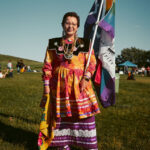 an indigenous person wearing traditional garments holding a 2spirit flag outside.